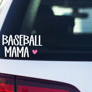 May include: White vinyl decal on a car window. The decal reads "BASEBALL MAMA" in a bold, stylized font, with a small pink heart symbol. The decal is placed on the rear window of a white car.
