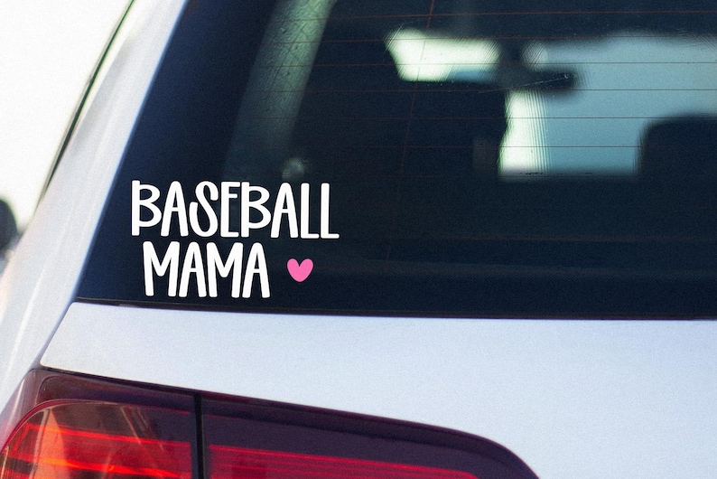 May include: White vinyl decal on a car window. The decal reads "BASEBALL MAMA" in a bold, stylized font, with a small pink heart symbol. The decal is placed on the rear window of a white car.