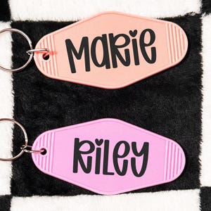 May include: Two retro-style keychains, one peach and one pink, each with a name in black script. The peach keychain says "Marie" with a heart, and the pink keychain says "Riley". Both have silver key rings.