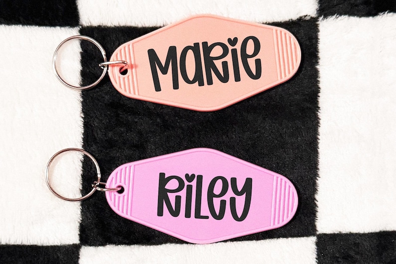 May include: Two retro-style keychains, one peach and one pink, each with a name in black script. The peach keychain says "Marie" with a heart, and the pink keychain says "Riley". Both have silver key rings.