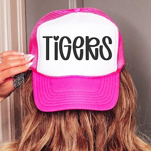 May include: A pink and white trucker hat with the word "TIGERS" in black script across the front. The hat has a mesh back and a curved brim. The hat is being held up by a person.