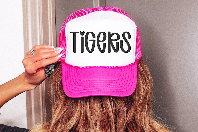May include: A pink and white trucker hat with the word "TIGERS" in black script across the front. The hat has a mesh back and a curved brim. The hat is being held up by a person.