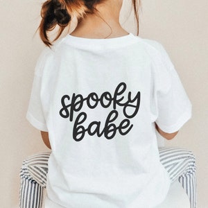 Puede incluir: Una camiseta blanca con las palabras "spooky babe" en escritura negra. La camiseta la lleva un ni&ntilde;o. El texto est&aacute; centrado en la parte posterior de la camiseta. El ni&ntilde;o est&aacute; sentado en una silla con tela a rayas.