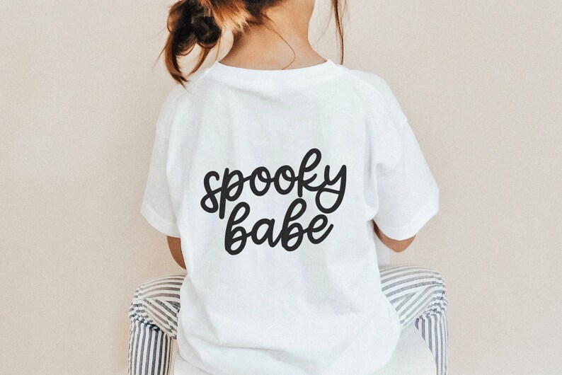 Puede incluir: Una camiseta blanca con las palabras "spooky babe" en escritura negra. La camiseta la lleva un ni&ntilde;o. El texto est&aacute; centrado en la parte posterior de la camiseta. El ni&ntilde;o est&aacute; sentado en una silla con tela a rayas.