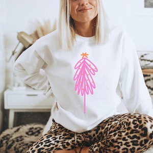May include: A white sweatshirt with a pink and orange abstract Christmas tree design. The tree has a star on top and is decorated with small orange circles.