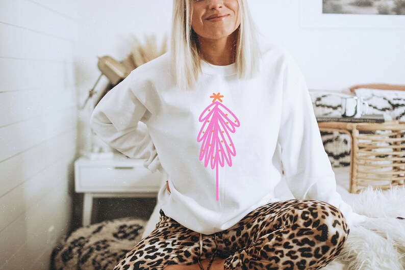 May include: A white sweatshirt with a pink and orange abstract Christmas tree design. The tree has a star on top and is decorated with small orange circles.