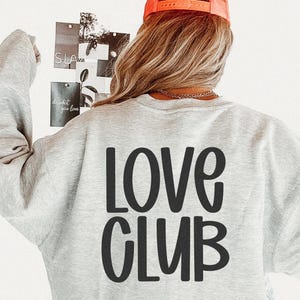 May include: A light gray sweatshirt with the words "LOVE CLUB" printed in large, black, rounded letters on the back. The person is wearing an orange baseball cap. The background includes a white wall and a collage of photos.