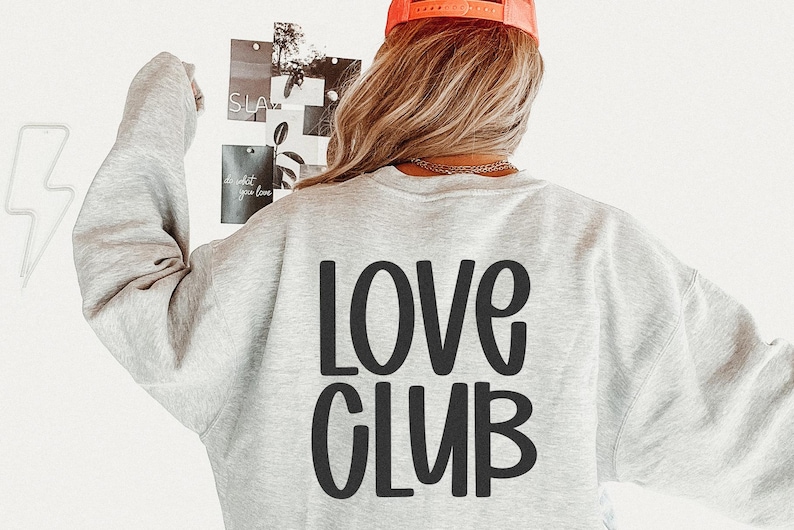 May include: A light gray sweatshirt with the words "LOVE CLUB" printed in large, black, rounded letters on the back. The person is wearing an orange baseball cap. The background includes a white wall and a collage of photos.