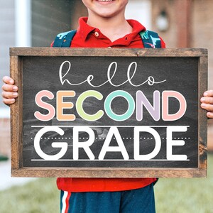 Op de afbeelding: Een kind houdt een bord van schoolbord met de tekst "hello second grade" in kleurrijke letters. Het bord is ingelijst met hout en heeft een zwart schoolbordoppervlak.