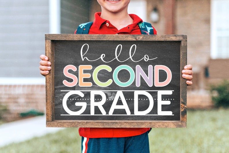 Op de afbeelding: Een kind houdt een bord van schoolbord met de tekst "hello second grade" in kleurrijke letters. Het bord is ingelijst met hout en heeft een zwart schoolbordoppervlak.