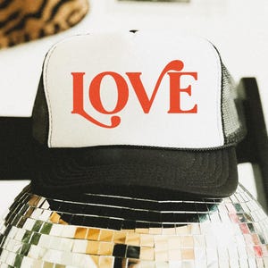 May include: A black and white trucker hat with the word "LOVE" in red script across the front. The hat is resting on a disco ball. The hat has a white front panel and a black mesh back.