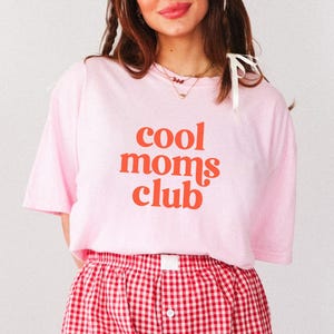 May include: A light pink t-shirt with the words "cool moms club" in orange lettering. The person is wearing red and white checkered shorts. The shirt is a casual, comfortable style.