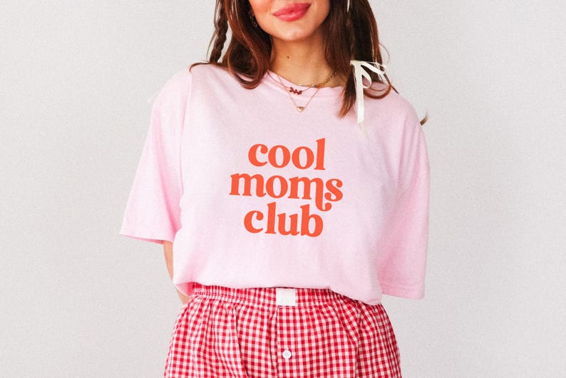 May include: A light pink t-shirt with the words "cool moms club" in orange lettering. The person is wearing red and white checkered shorts. The shirt is a casual, comfortable style.