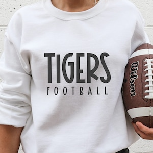 May include: White sweatshirt with black text that reads "TIGERS FOOTBALL". A person is holding a brown and white football with the word "Wilson" printed on it.