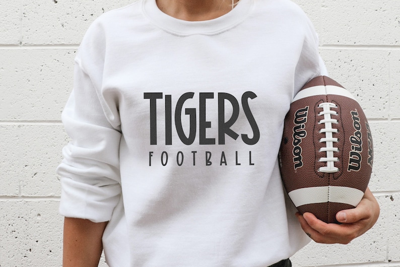 May include: White sweatshirt with black text that reads "TIGERS FOOTBALL". A person is holding a brown and white football with the word "Wilson" printed on it.