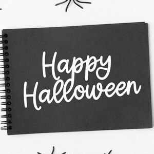 Puede incluir: Un cuaderno negro con espiral con las palabras "Happy Halloween" en escritura blanca. Ara&ntilde;as de pl&aacute;stico negro est&aacute;n esparcidas alrededor del cuaderno sobre un fondo blanco. La imagen tiene un tema festivo y de temporada.