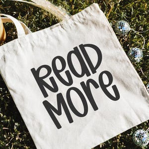 May include: A beige tote bag with the words "READ MORE" in bold, black letters. The bag is on a bed of green grass, with a string of small disco ball lights and a glass tumbler with a wooden lid.