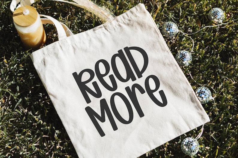 May include: A beige tote bag with the words "READ MORE" in bold, black letters. The bag is on a bed of green grass, with a string of small disco ball lights and a glass tumbler with a wooden lid.