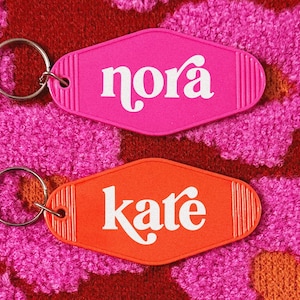May include: Two retro-style keychains, one pink and one orange, each with a silver keyring. The pink keychain reads "nora" in white script, and the orange keychain reads "kate". The background is a textured, floral pattern.