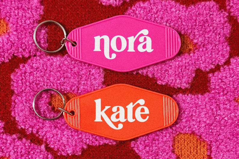 May include: Two retro-style keychains, one pink and one orange, each with a silver keyring. The pink keychain reads "nora" in white script, and the orange keychain reads "kate". The background is a textured, floral pattern.