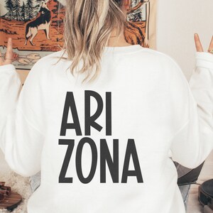 May include: White sweatshirt with black text that reads "ARI ZONA" on the back. The person wearing the sweatshirt is making the peace sign with both hands.