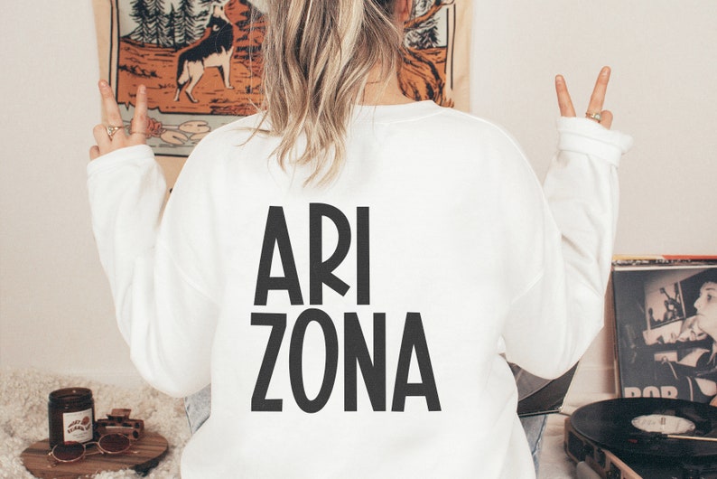 May include: White sweatshirt with black text that reads "ARI ZONA" on the back. The person wearing the sweatshirt is making the peace sign with both hands.