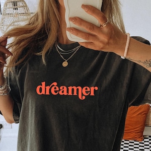 May include: Dark gray t-shirt with the word "dreamer" in red script. The shirt is a relaxed fit and appears to be made of a soft material. The image shows the shirt being worn, with jewelry and a tattoo visible.