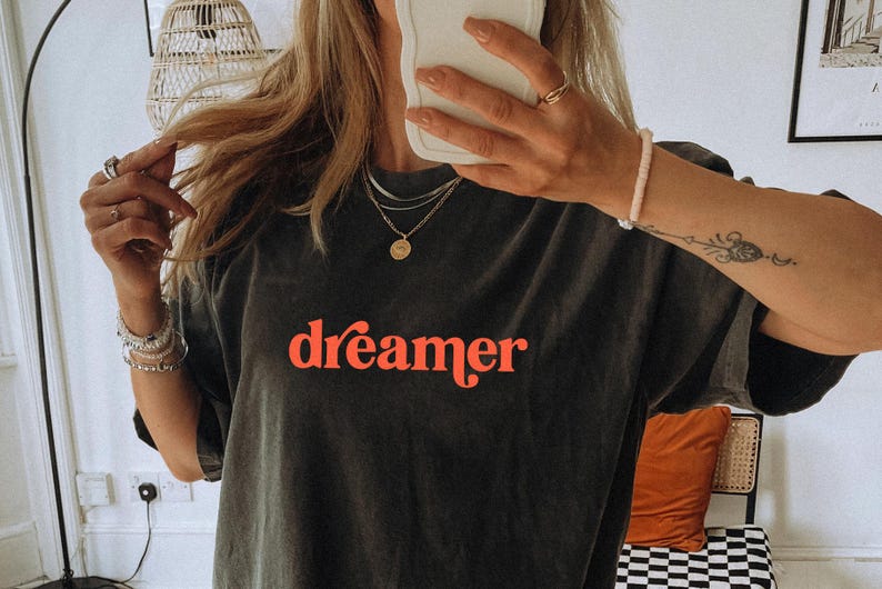 May include: Dark gray t-shirt with the word "dreamer" in red script. The shirt is a relaxed fit and appears to be made of a soft material. The image shows the shirt being worn, with jewelry and a tattoo visible.