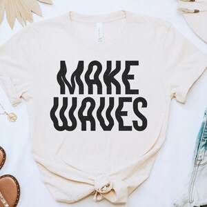 Op de afbeelding: Een wit T-shirt met de tekst "MAKE WAVES" in zwarte, golvende letters. Het shirt ligt plat op een wit oppervlak met andere accessoires.
