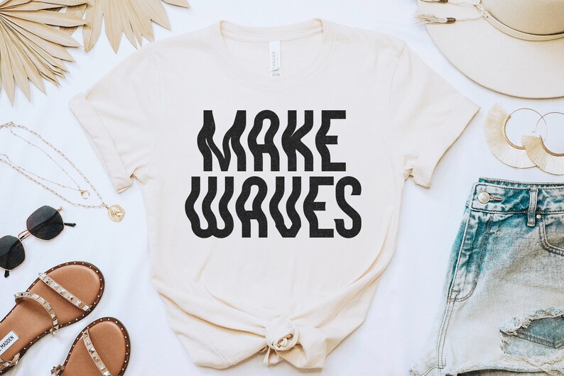 Op de afbeelding: Een wit T-shirt met de tekst "MAKE WAVES" in zwarte, golvende letters. Het shirt ligt plat op een wit oppervlak met andere accessoires.