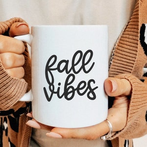 Puede incluir: Taza de cer&aacute;mica blanca con las palabras "fall vibes" en escritura negra. La taza la sostiene una persona que lleva un su&eacute;ter marr&oacute;n con un dise&ntilde;o de estampado de leopardo.