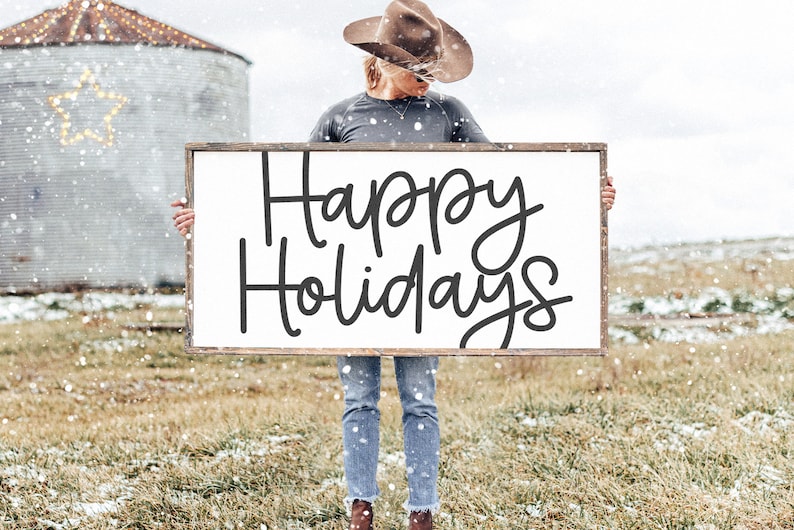 Peut inclure: Une personne tenant une pancarte blanche avec une inscription noire en cursive qui dit "Happy Holidays". La pancarte est encadr&eacute;e de bois et la personne porte un chapeau de cowboy marron et un jean bleu. L'arri&egrave;re-plan est un champ enneig&eacute;.