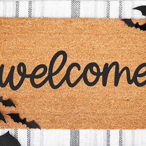 Puede incluir: Una alfombra rectangular con la palabra "welcome" en escritura negra. La alfombra est&aacute; hecha de un material marr&oacute;n claro y est&aacute; decorada con recortes de murci&eacute;lagos negros en los bordes. El fondo es un patr&oacute;n de cuadros blancos y negros.