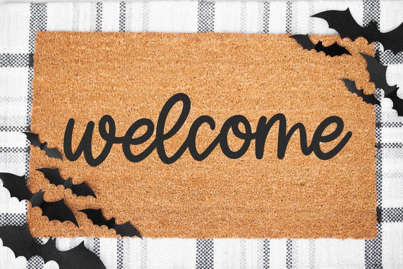 Puede incluir: Una alfombra rectangular con la palabra "welcome" en escritura negra. La alfombra est&aacute; hecha de un material marr&oacute;n claro y est&aacute; decorada con recortes de murci&eacute;lagos negros en los bordes. El fondo es un patr&oacute;n de cuadros blancos y negros.