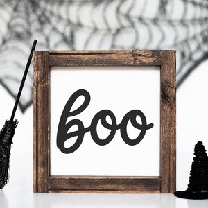 Puede incluir: Un letrero de madera con tem&aacute;tica de Halloween con la palabra "boo" en escritura negra. El letrero est&aacute; enmarcado en madera marr&oacute;n oscuro. Un sombrero de bruja negro y una escoba negra se colocan cerca del letrero, con una telara&ntilde;a en el fondo.
