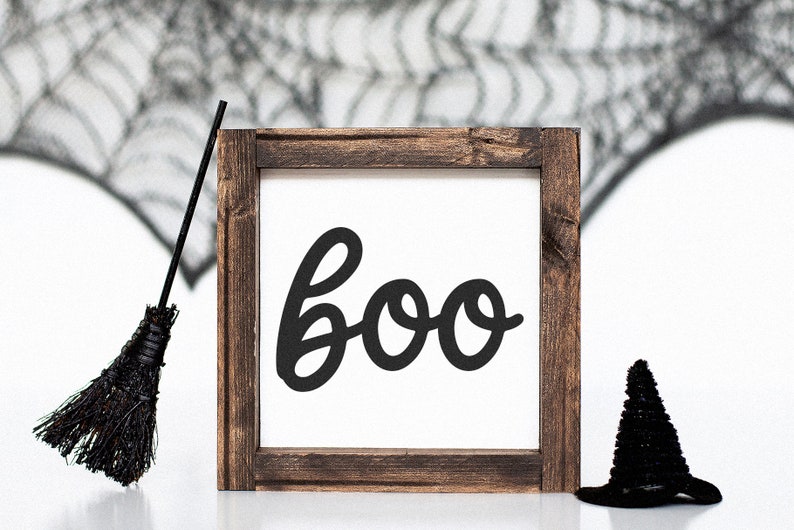 Puede incluir: Un letrero de madera con tem&aacute;tica de Halloween con la palabra "boo" en escritura negra. El letrero est&aacute; enmarcado en madera marr&oacute;n oscuro. Un sombrero de bruja negro y una escoba negra se colocan cerca del letrero, con una telara&ntilde;a en el fondo.