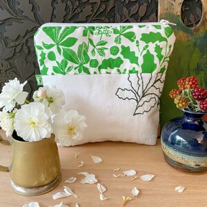 May include: A white and green floral print zippered pouch with a white base and a black leaf design. A brass mug with white flowers and a small blue vase with berries are in the foreground.
