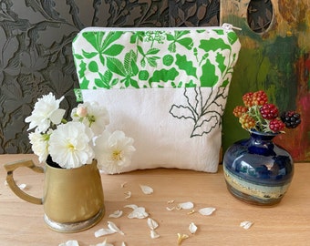 May include: A white and green floral print zippered pouch with a white base and a black leaf design. A brass mug with white flowers and a small blue vase with berries are in the foreground.