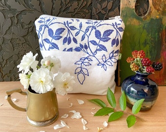 May include: A white fabric pouch with a blue floral design and a zipper closure. The pouch is sitting on a wooden table with a small brass pitcher and a blue ceramic vase with green leaves and berries.