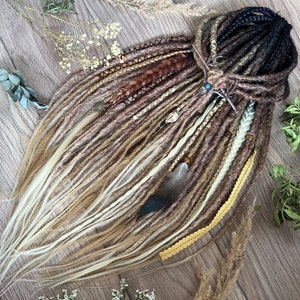 Op de afbeelding: Een set van bruine, blonde en zwarte synthetische dreadlocks met kralen, veren en een gouden ketting. De dreadlocks zijn in een waaier op een houten oppervlak gerangschikt.