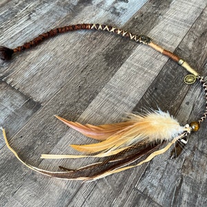 Op de afbeelding: Een gevlochten haarverlenging in bruin en beige met een bruine veer, een bruin en beige lint en een gouden bedel. De verlenging is vastgebonden met een bruin touwtje.