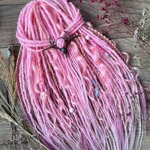 May include: Pink and purple synthetic dreadlocks with decorative beads and charms. The dreadlocks are styled with a dark metal antler hair accessory. The hair is arranged on a wooden surface with dried flowers.