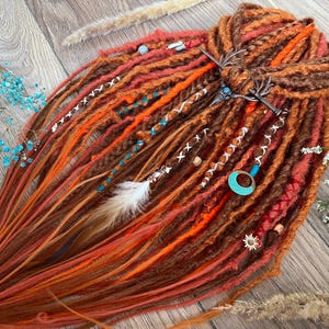 Puede incluir: Un conjunto de rastas sintéticas largas en tonos naranja, marrón y rojo. Las rastas están decoradas con cuentas, plumas y una pinza para el pelo de metal.