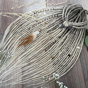 Op de afbeelding: Een set lange, blonde synthetische dreadlocks met bruine en witte kralen, bruin en wit touw en een bruine verenaccent.