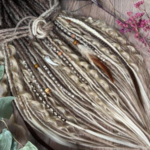 Puede incluir: Dreadlocks sintéticas marrones y rubias con cuentas, plumas y un amuleto de hueso. Las dreadlocks están dispuestas en cascada.