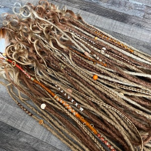 Könnte beinhalten: Ein Set aus braunen, blonden und beigefarbenen synthetischen Dreadlocks mit verschiedenen Perlen und Charms. Die Dreadlocks sind fächerförmig auf einer Holzoberfläche angeordnet.