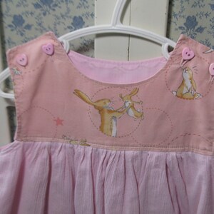 May include: A pink sleeveless dress with a pink and white fabric top featuring a bunny rabbit design. The dress has a gathered skirt and two pink heart-shaped buttons on the straps.