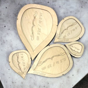 Könnte beinhalten: Fünf herzförmige Holzausschnitte mit Maßen in Zoll und Zentimetern. Der größte Ausschnitt ist 14,8 Zoll mal 37,6 Zentimeter groß. Der kleinste Ausschnitt ist 5 Zoll mal 13 Zentimeter groß. Die Ausschnitte sind mit "Cordas" und "MB" beschriftet.