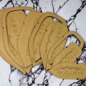 Può includere: Un set di sei modelli di asse da stiro in legno marrone con misure in pollici e centimetri. I modelli hanno la forma di un asse da stiro e hanno un design decorativo.