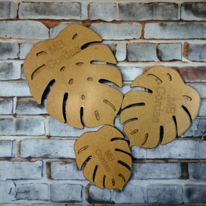 Peut inclure: Trois formes de feuilles de monstera en bois avec le texte "MB Cordas" sur chacune. Les feuilles sont de tailles différentes et sont disposées sur un fond de mur en briques.